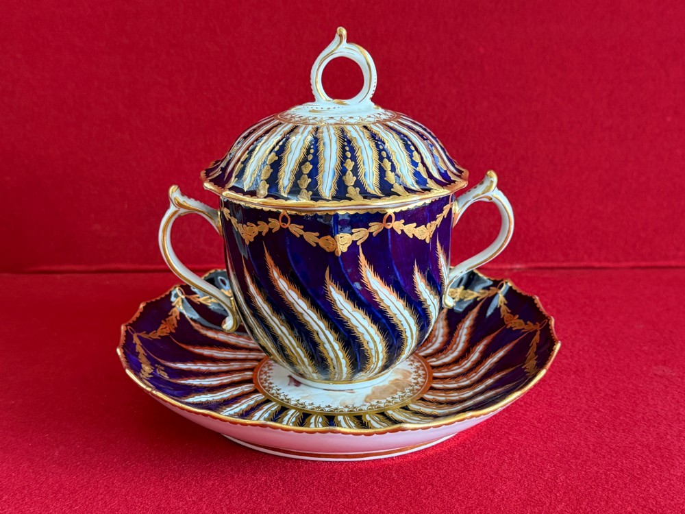 An 18th-century cobalt blue and gilded soft-paste porcelain chocolate cup with lid alongside a matching saucer decorated with a painted central cherub motif. View II