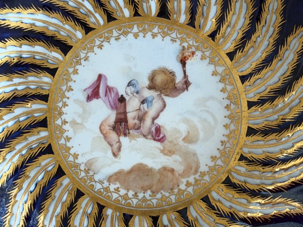 An 18th-century cobalt blue and gilded soft-paste porcelain chocolate cup with lid alongside a matching saucer decorated with a painted central cherub motif. View IV