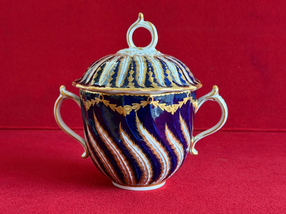 An 18th-century cobalt blue and gilded soft-paste porcelain chocolate cup with lid alongside a matching saucer decorated with a painted central cherub motif. View VIII
