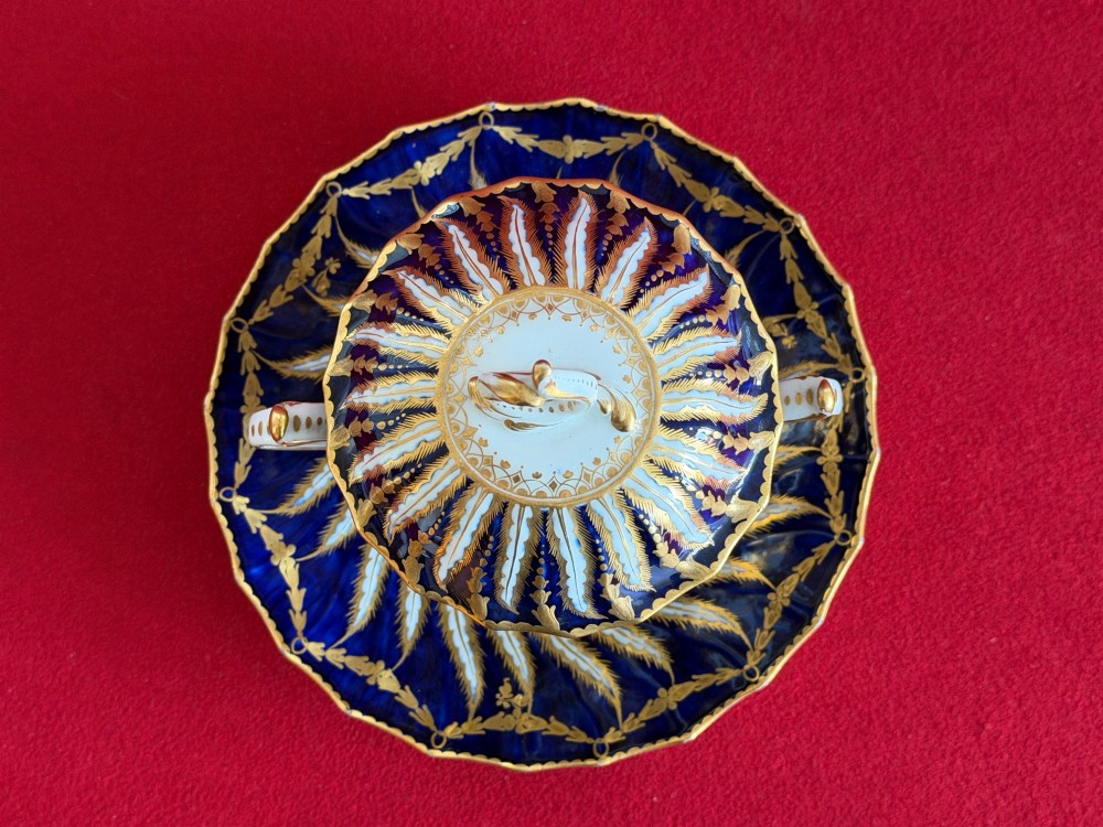An 18th-century cobalt blue and gilded soft-paste porcelain chocolate cup with lid alongside a matching saucer decorated with a painted central cherub motif. View X