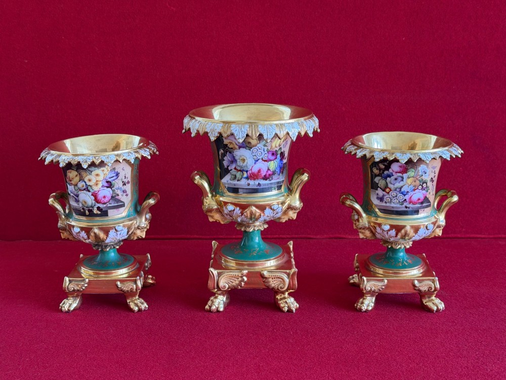 A three-piece antique porcelain garniture set featuring green and gold gilded urns with hand-painted floral panels on pedestal bases with paw feet.