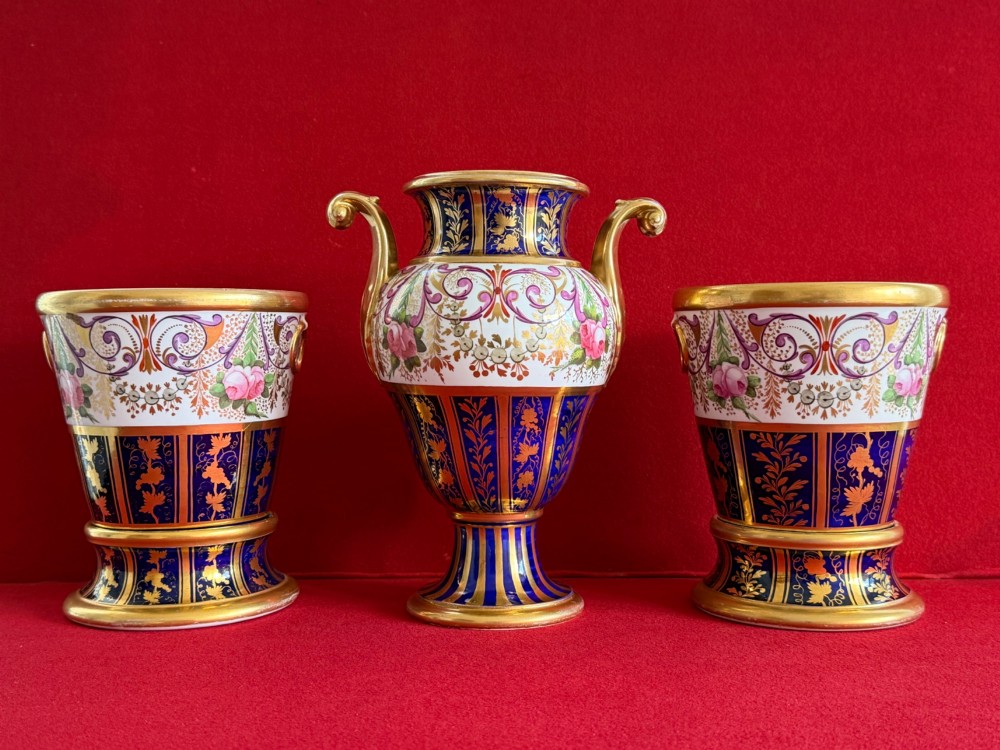 A three-piece English Regency Spode porcelain garniture featuring a central twin-handled urn flanked by two beaker-shaped vases decorated with cobalt blue panels, hand-painted pink floral motifs, and detailed gilt scrollwork.