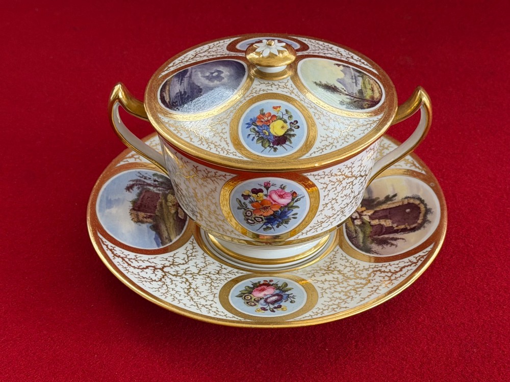 An antique Flight, Barr and Barr Worcester porcelain covered chocolate cup with two handles and matching saucer, decorated with hand-painted landscape and floral panels amid gold vermicelli detailing.
