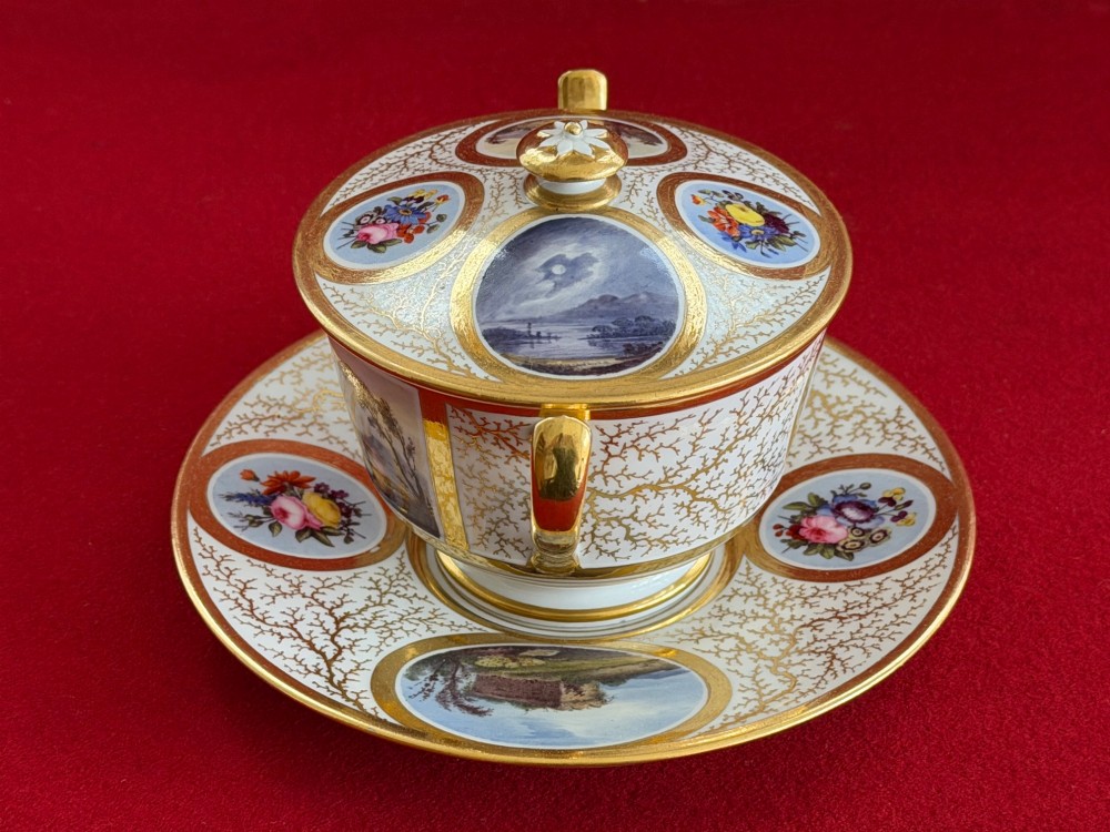An antique Flight, Barr and Barr Worcester porcelain covered chocolate cup with two handles and matching saucer, decorated with hand-painted landscape and floral panels amid gold vermicelli detailing. View II