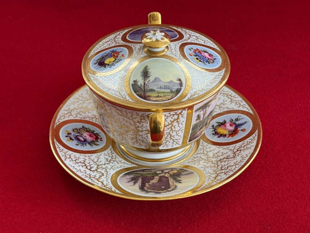 An antique Flight, Barr and Barr Worcester porcelain covered chocolate cup with two handles and matching saucer, decorated with hand-painted landscape and floral panels amid gold vermicelli detailing. View IV