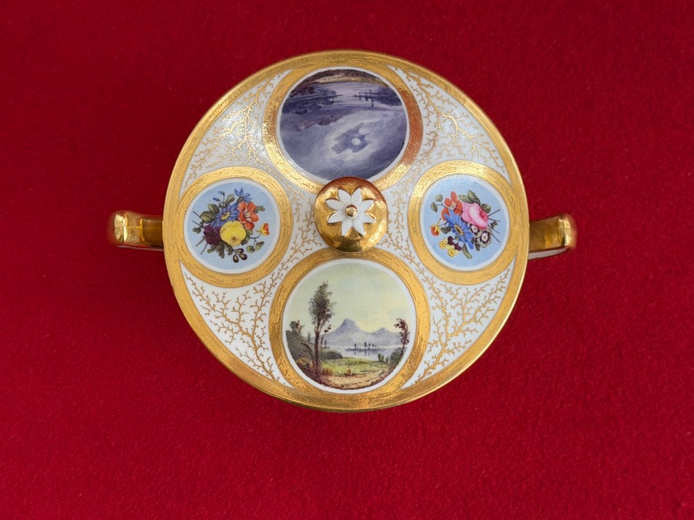 An antique Flight, Barr and Barr Worcester porcelain covered chocolate cup with two handles and matching saucer, decorated with hand-painted landscape and floral panels amid gold vermicelli detailing. View VIII