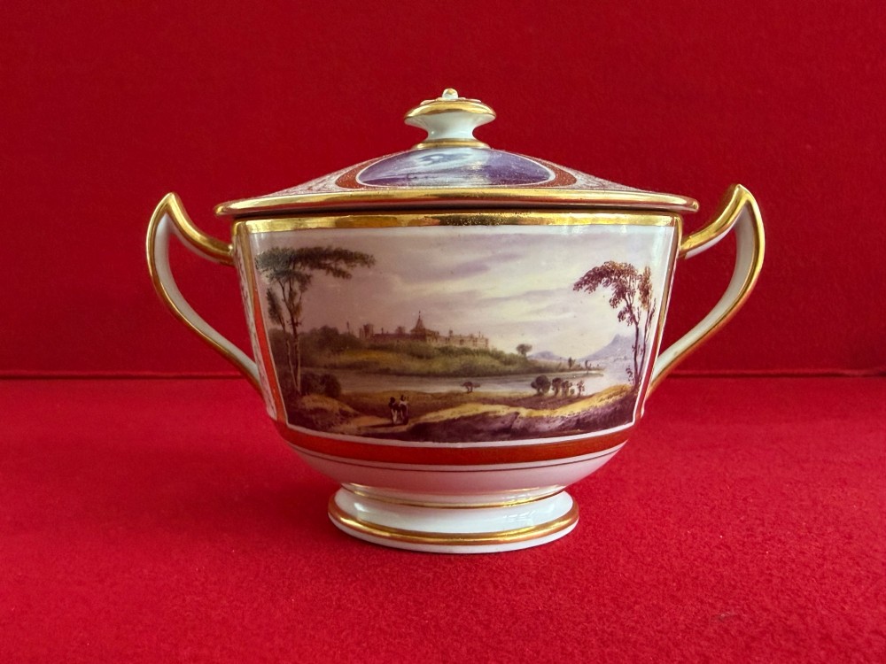 An antique Flight, Barr and Barr Worcester porcelain covered chocolate cup with two handles and matching saucer, decorated with hand-painted landscape and floral panels amid gold vermicelli detailing. View X