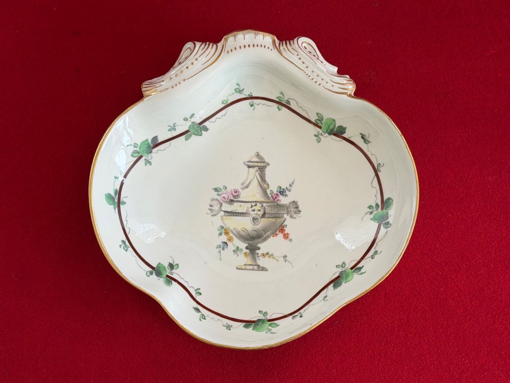 An 18th-century shell-shaped white soft-paste porcelain dish decorated with a central classical urn motif, green leafy garland border, and gilded rim details.