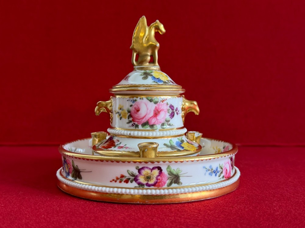 A circular white porcelain inkstand featuring hand-painted floral decoration, gilt accents, pen holders, and a cover topped with a golden griffin finial. View VI
