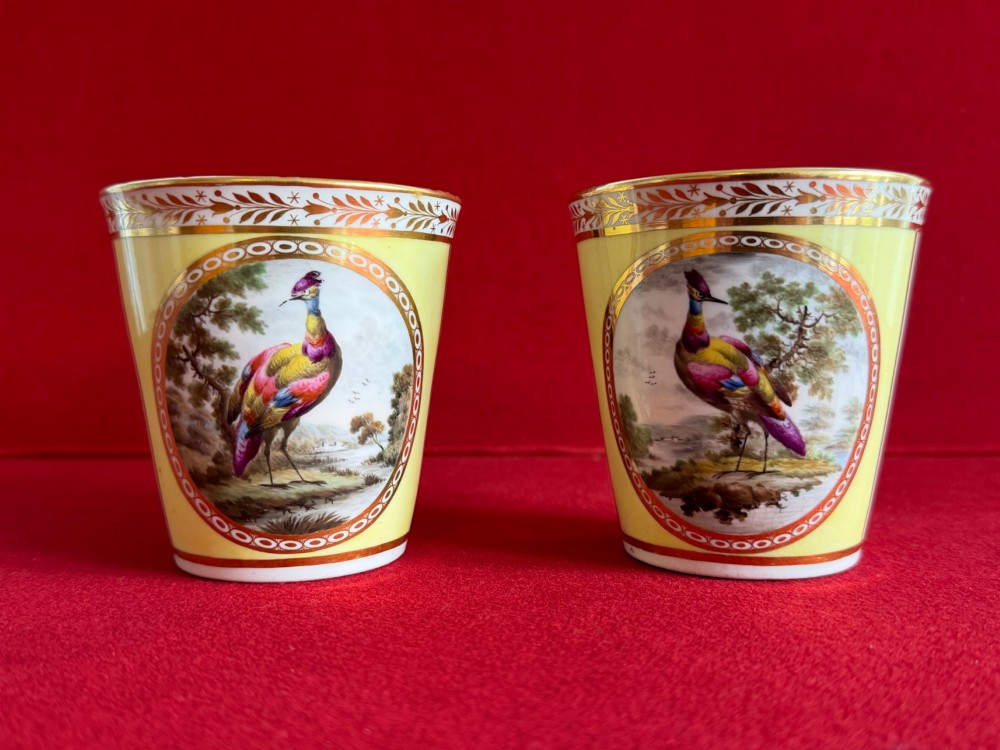 A pair of yellow antique porcelain beakers featuring hand-painted exotic birds within oval gilt reserves and gilded leaf borders along the top rim.