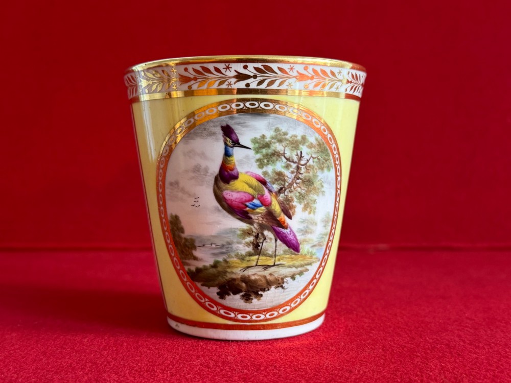 A pair of yellow antique porcelain beakers featuring hand-painted exotic birds within oval gilt reserves and gilded leaf borders along the top rim. View V