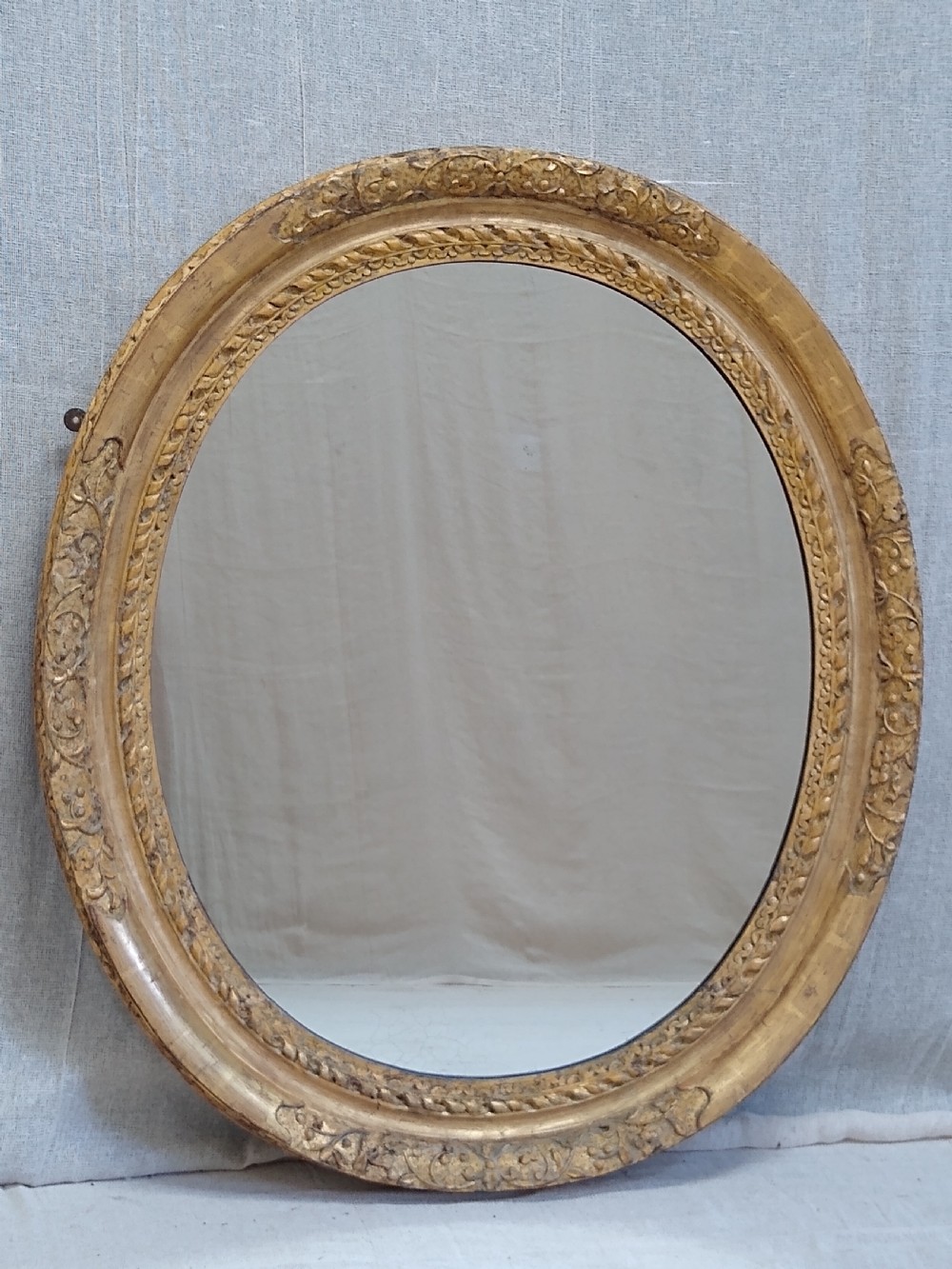 An antique oval wall mirror featuring a heavily hand-carved giltwood frame decorated with foliate and leaf moulding. View IV