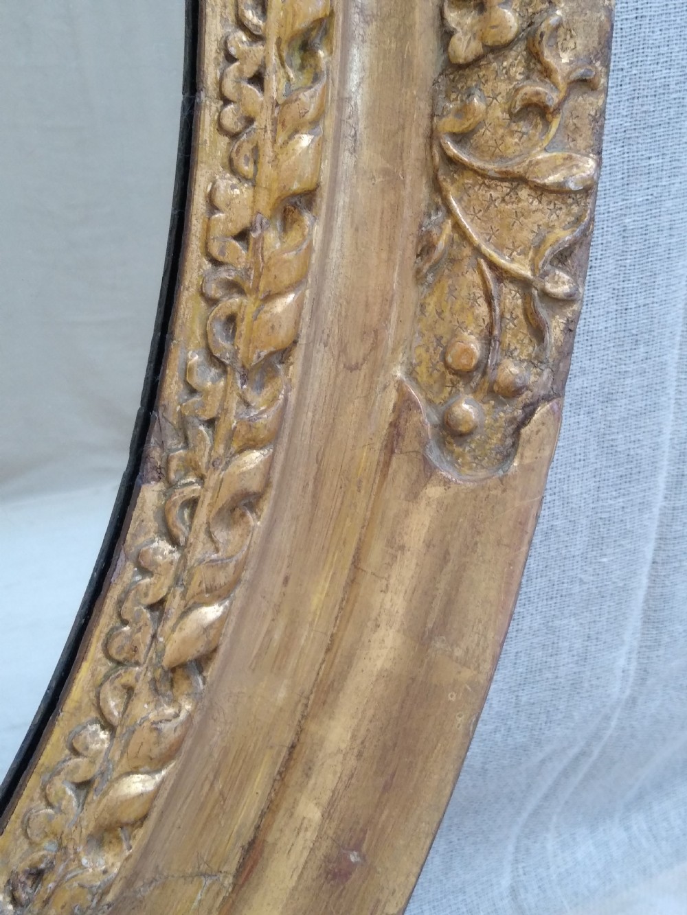 An antique oval wall mirror featuring a heavily hand-carved giltwood frame decorated with foliate and leaf moulding. View V