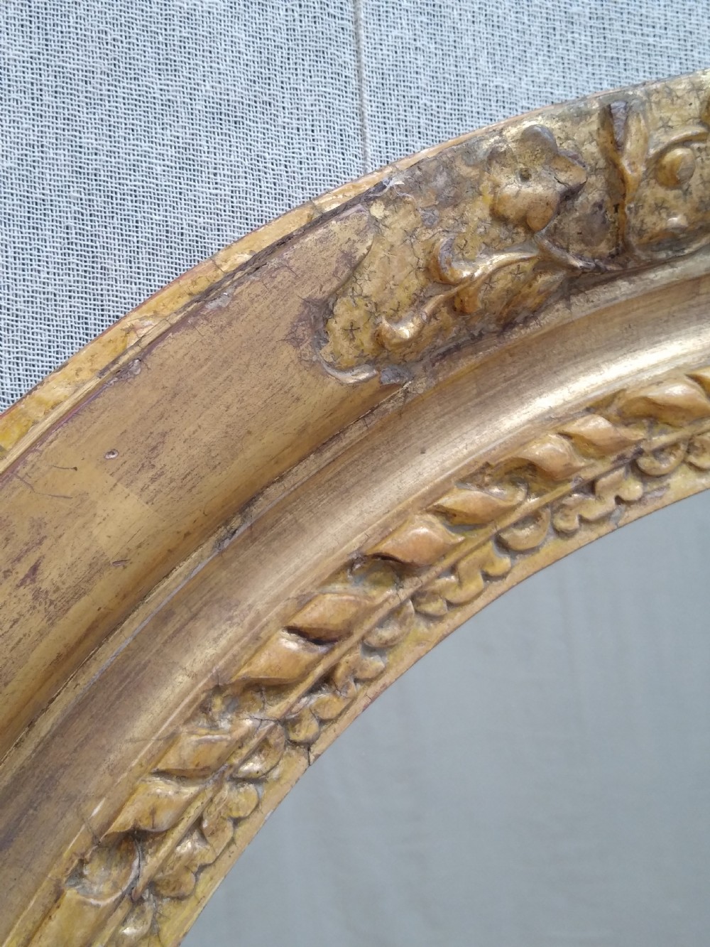 An antique oval wall mirror featuring a heavily hand-carved giltwood frame decorated with foliate and leaf moulding. View IX