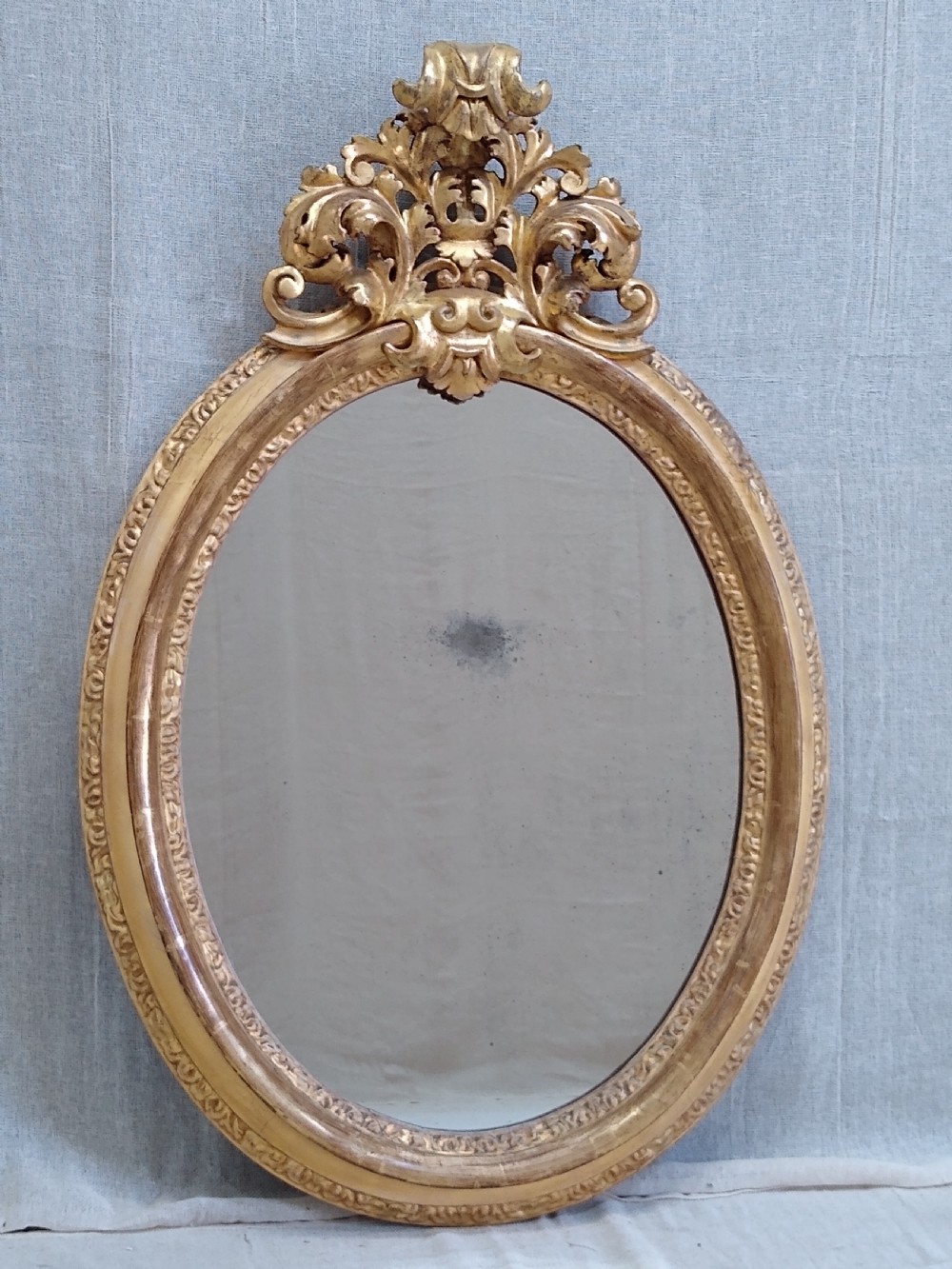 An antique 19th-century Italian oval giltwood wall mirror with an ornate carved scroll cresting pediment. View III