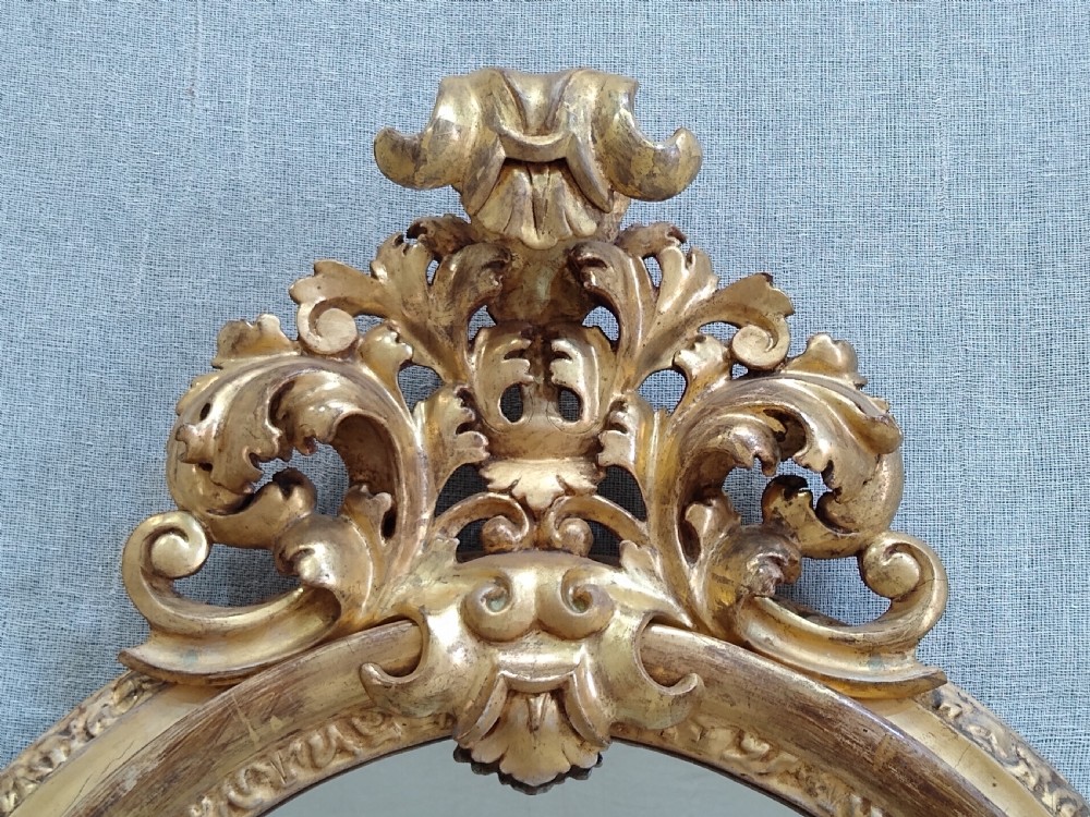 An antique 19th-century Italian oval giltwood wall mirror with an ornate carved scroll cresting pediment. View VII