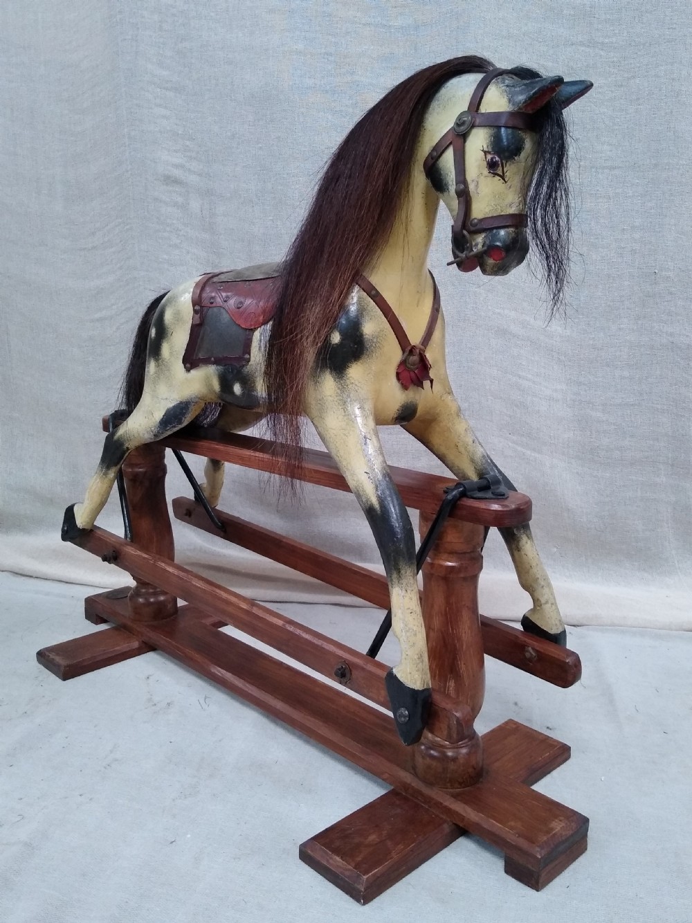 An antique carved wooden rocking horse with a painted dapple finish, leather saddle, and real horsehair mane, mounted on a wooden safety stand.