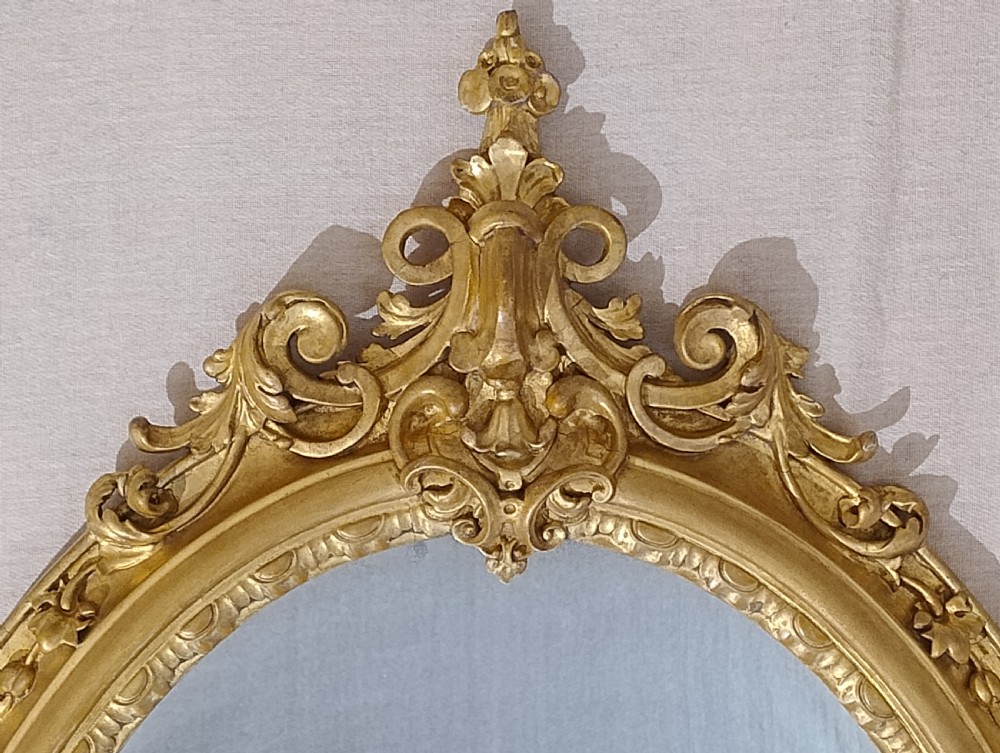 An antique Victorian oval wall mirror featuring an ornate gilded frame with carved scrollwork cresting and apron. View III