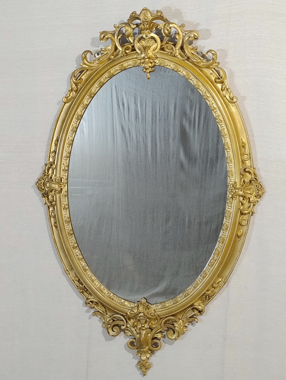 An antique Victorian oval wall mirror featuring an ornate gilded frame with carved scrollwork cresting and apron.