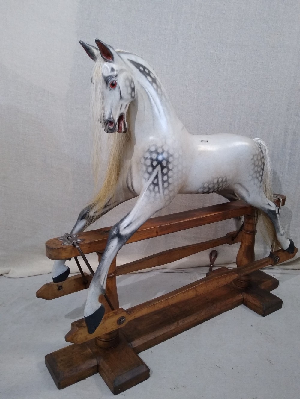 An antique carved wooden rocking horse with a dapple grey painted finish, real hair mane, leather saddle, and bridle, mounted on a wooden safety stand. View IV