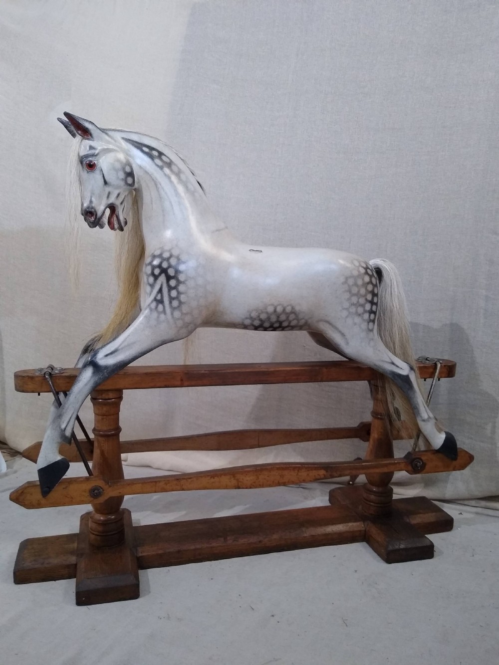 An antique carved wooden rocking horse with a dapple grey painted finish, real hair mane, leather saddle, and bridle, mounted on a wooden safety stand. View III