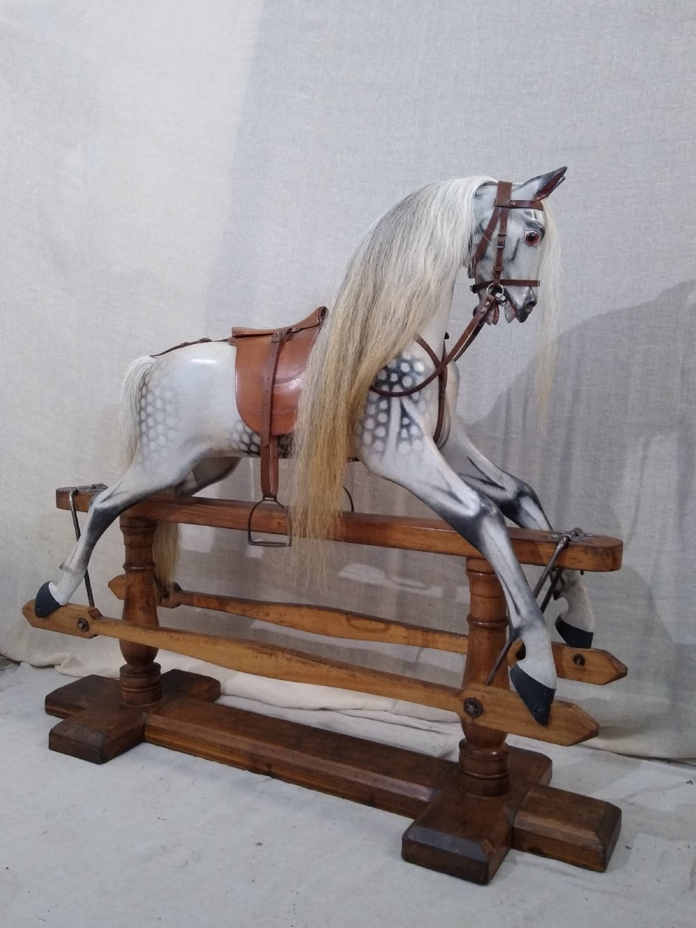 An antique carved wooden rocking horse with a dapple grey painted finish, real hair mane, leather saddle, and bridle, mounted on a wooden safety stand.