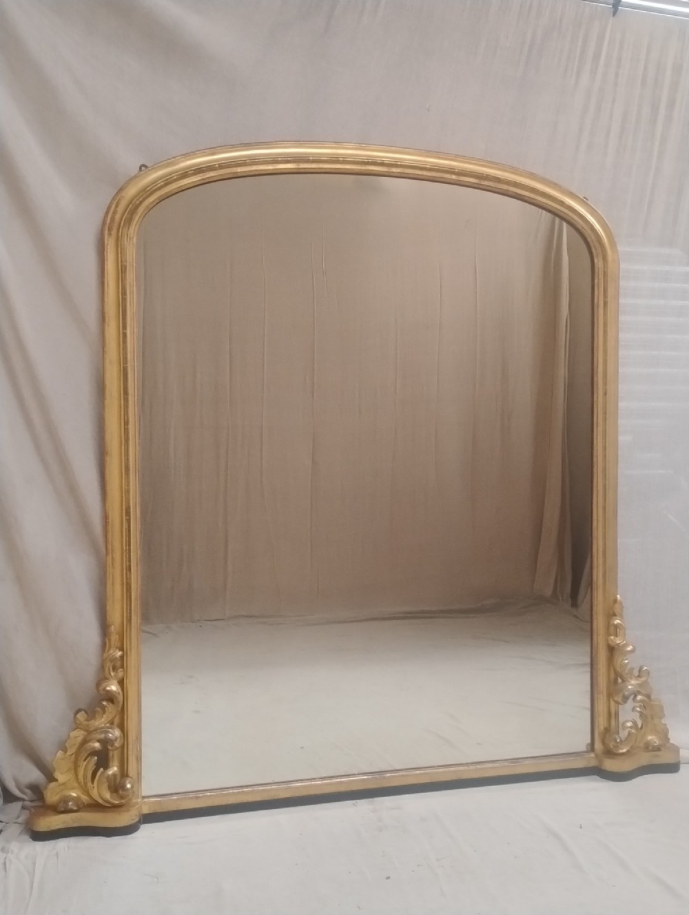 A large arched antique overmantel mirror featuring a gold giltwood frame with carved acanthus leaf scrolls at the lower corners.