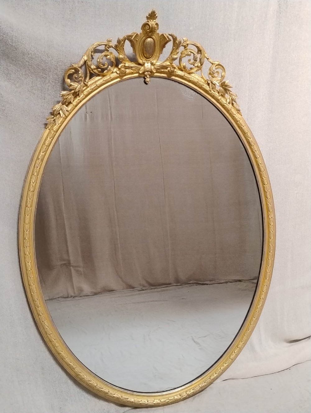 An antique oval wall mirror featuring a giltwood frame topped with an ornate leaf cresting and enclosing a mercury glass plate.