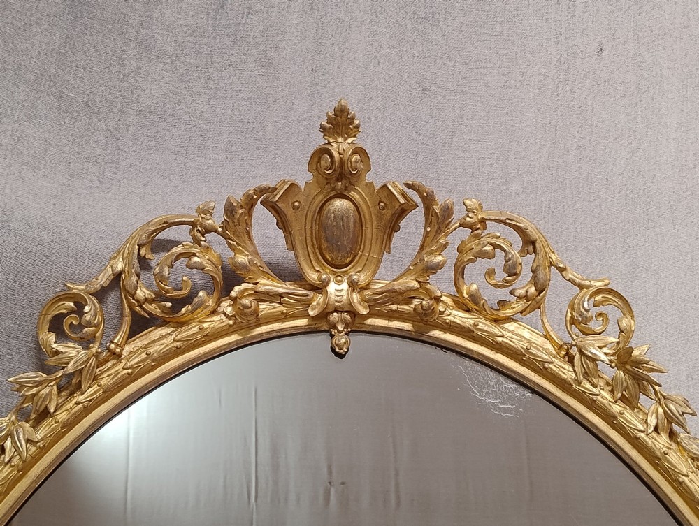 An antique oval wall mirror featuring a giltwood frame topped with an ornate leaf cresting and enclosing a mercury glass plate. View IV