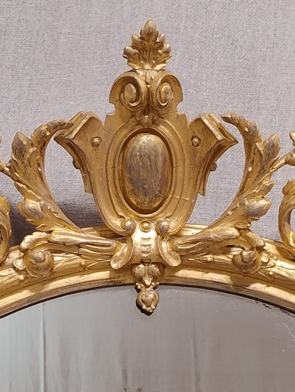 An antique oval wall mirror featuring a giltwood frame topped with an ornate leaf cresting and enclosing a mercury glass plate. View V