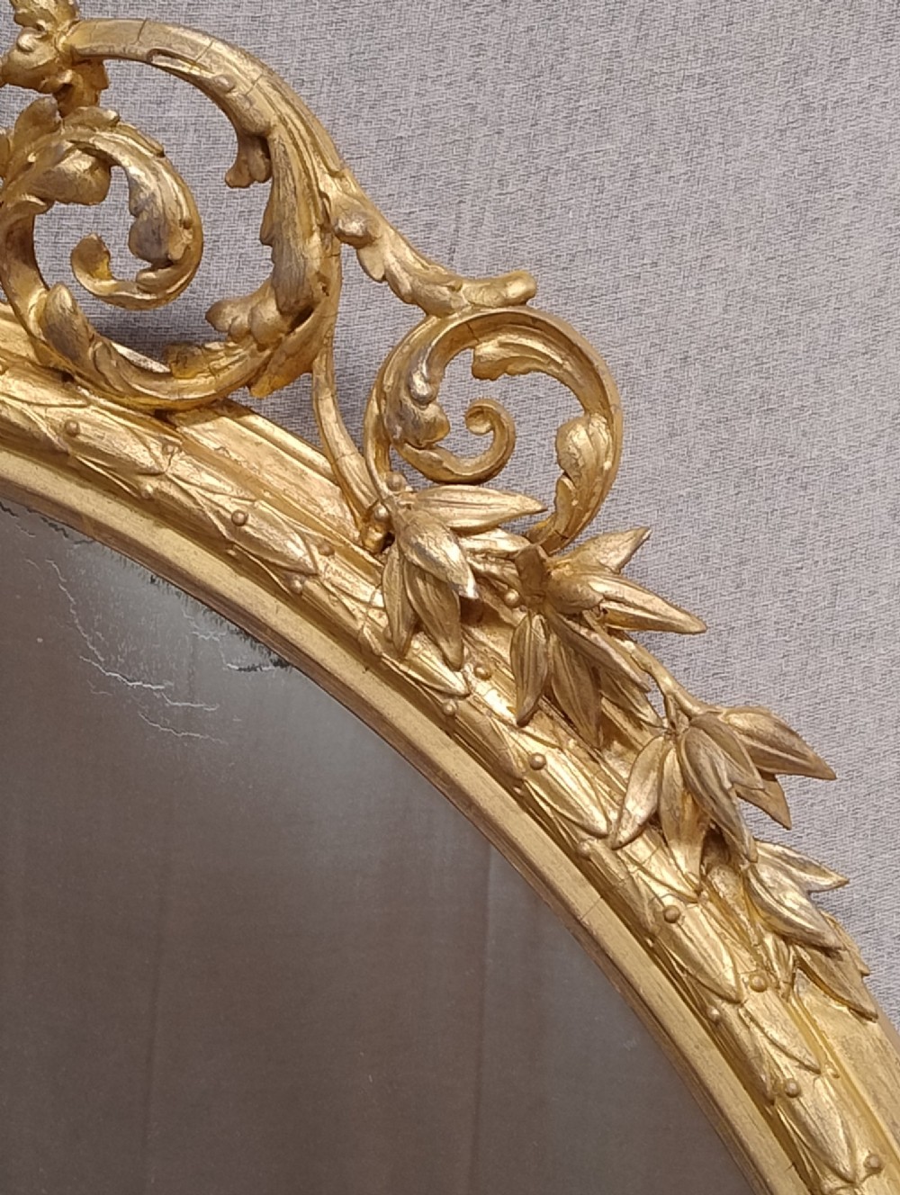 An antique oval wall mirror featuring a giltwood frame topped with an ornate leaf cresting and enclosing a mercury glass plate. View VI