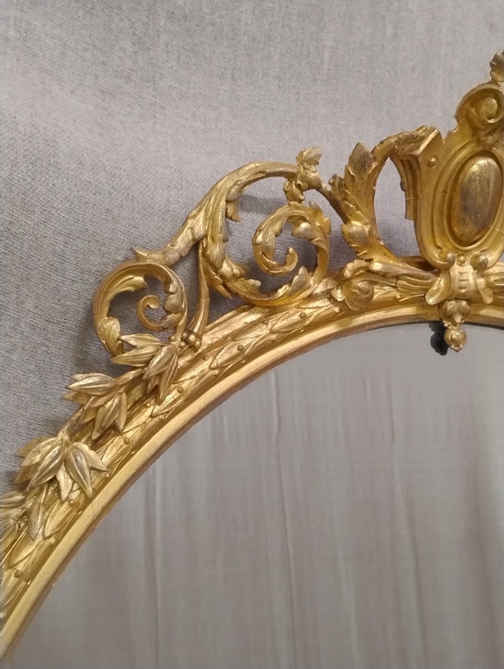An antique oval wall mirror featuring a giltwood frame topped with an ornate leaf cresting and enclosing a mercury glass plate. View VII