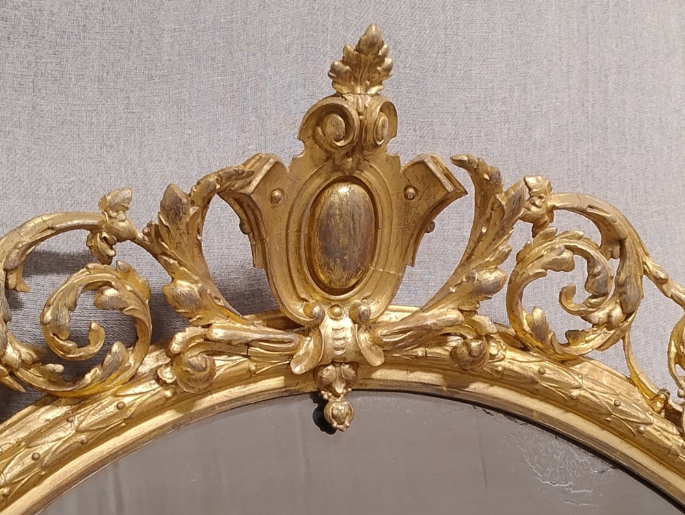 An antique oval wall mirror featuring a giltwood frame topped with an ornate leaf cresting and enclosing a mercury glass plate. View VIII