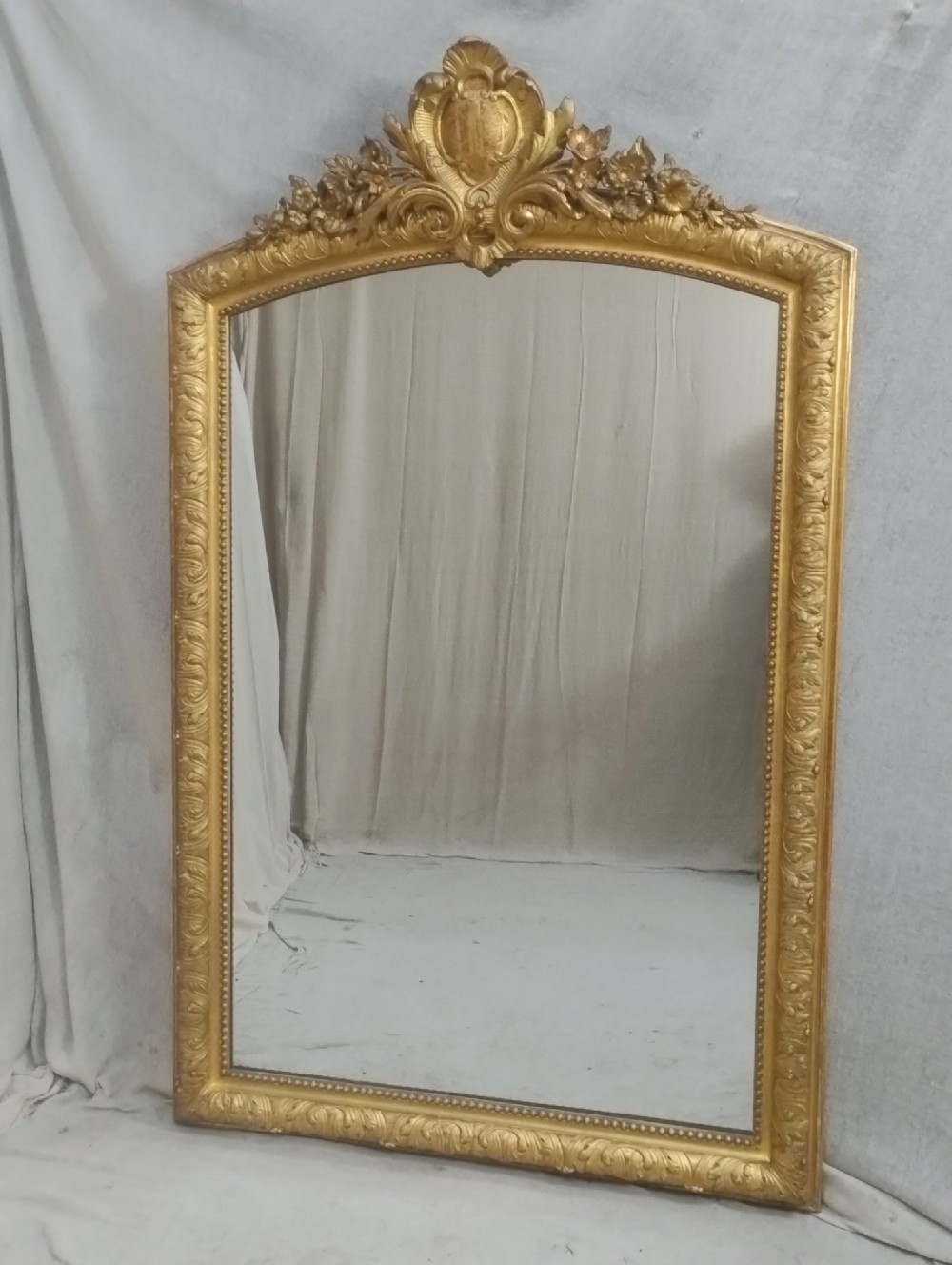 A tall French giltwood mirror featuring a carved floral cartouche cresting, arched top, and moulded beaded inner border.