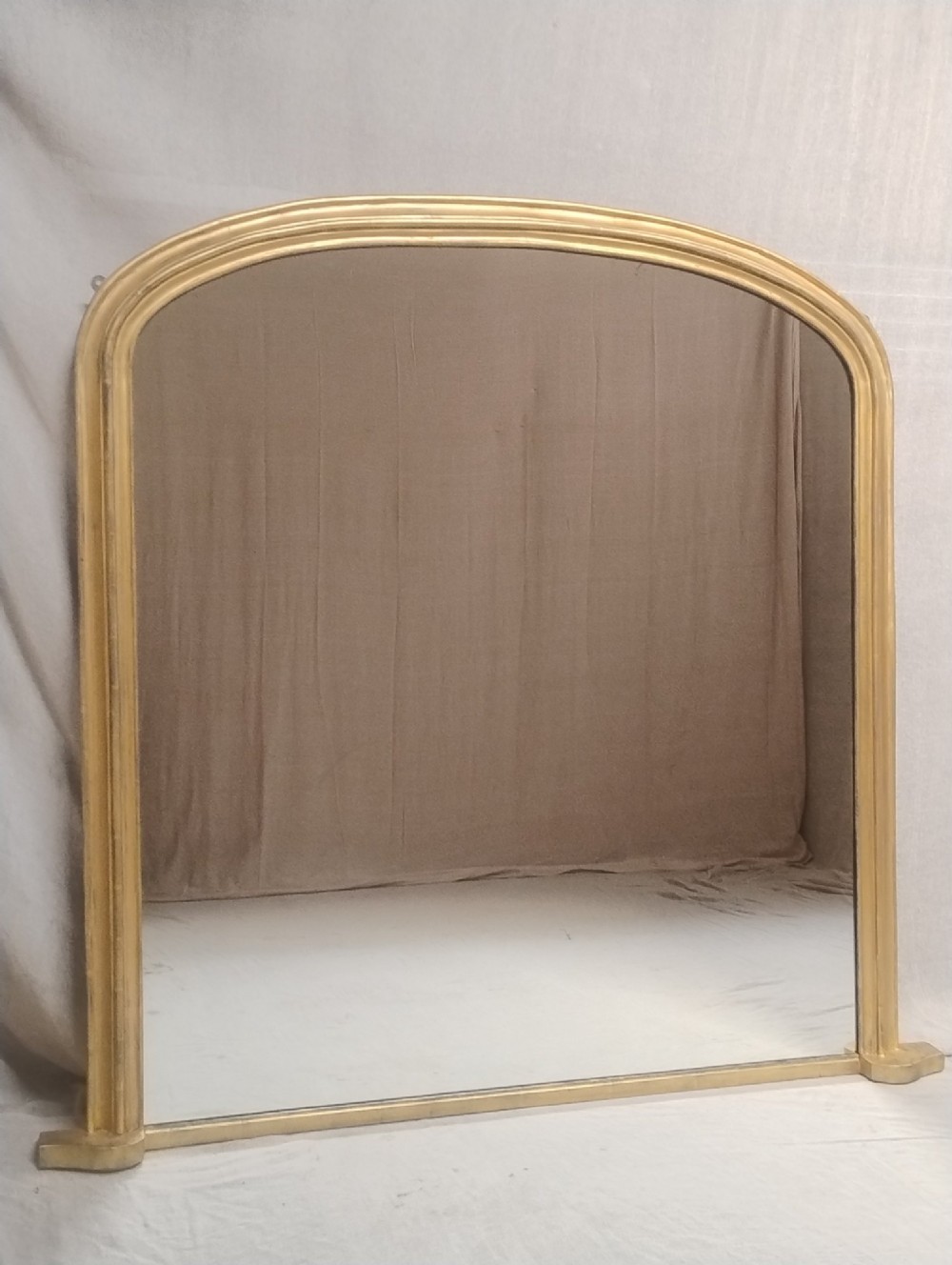 An antique Victorian arched overmantel mirror featuring a moulded gold giltwood frame with subtle bottom side corbels.