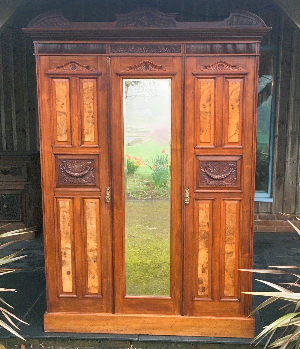 A late Victorian double wardrobe in warm brown walnut featuring a central full-length mirrored door flanked by two carved and burr walnut panelled doors beneath a moulded pediment.