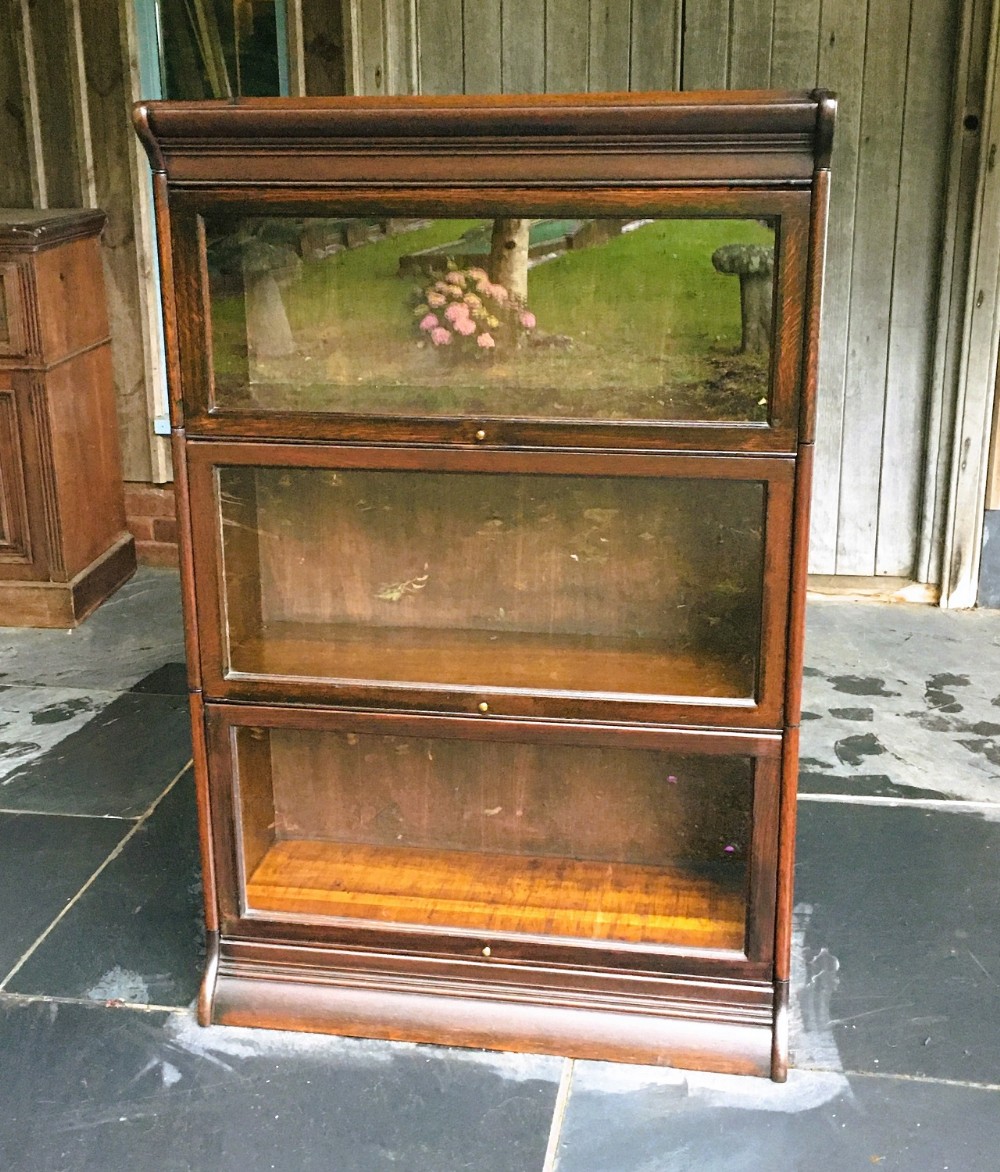 A three-tier dark oak sectional bookcase with glazed lift-up doors and small brass knobs on a moulded base.