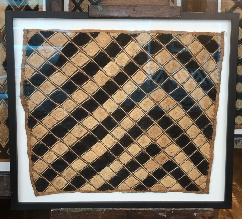 A framed geometric Kuba raffia textile featuring a diagonal grid pattern in natural tan and dark brown tones, presented within a black wooden frame and white mount.