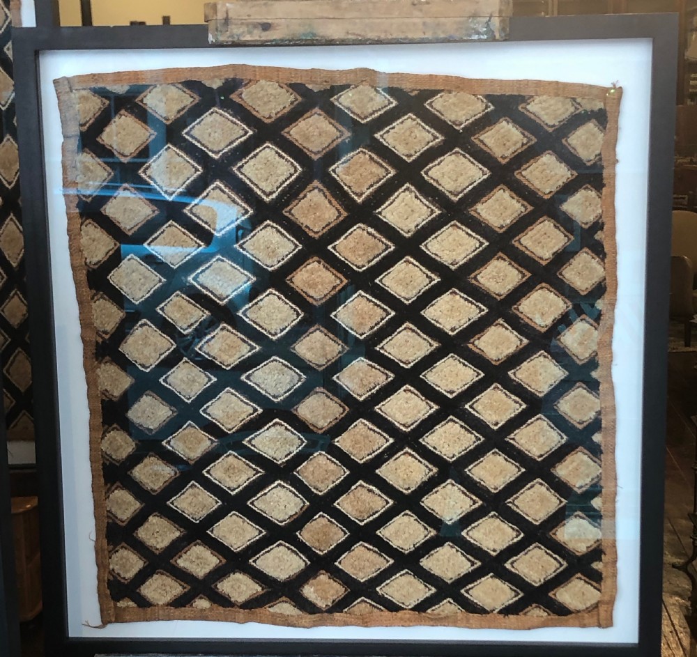 A framed geometric Kuba raffia textile featuring a diagonal grid pattern in natural tan and dark brown tones, presented within a black wooden frame and white mount. View II