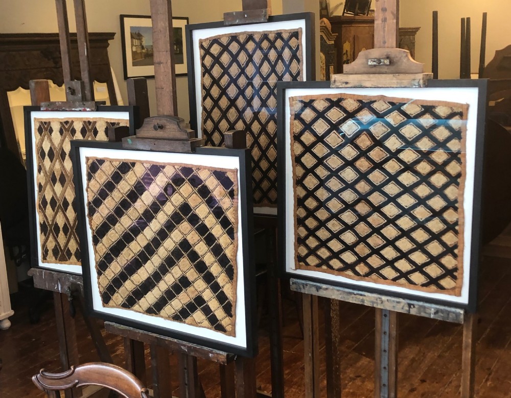 A framed geometric Kuba raffia textile featuring a diagonal grid pattern in natural tan and dark brown tones, presented within a black wooden frame and white mount. View III
