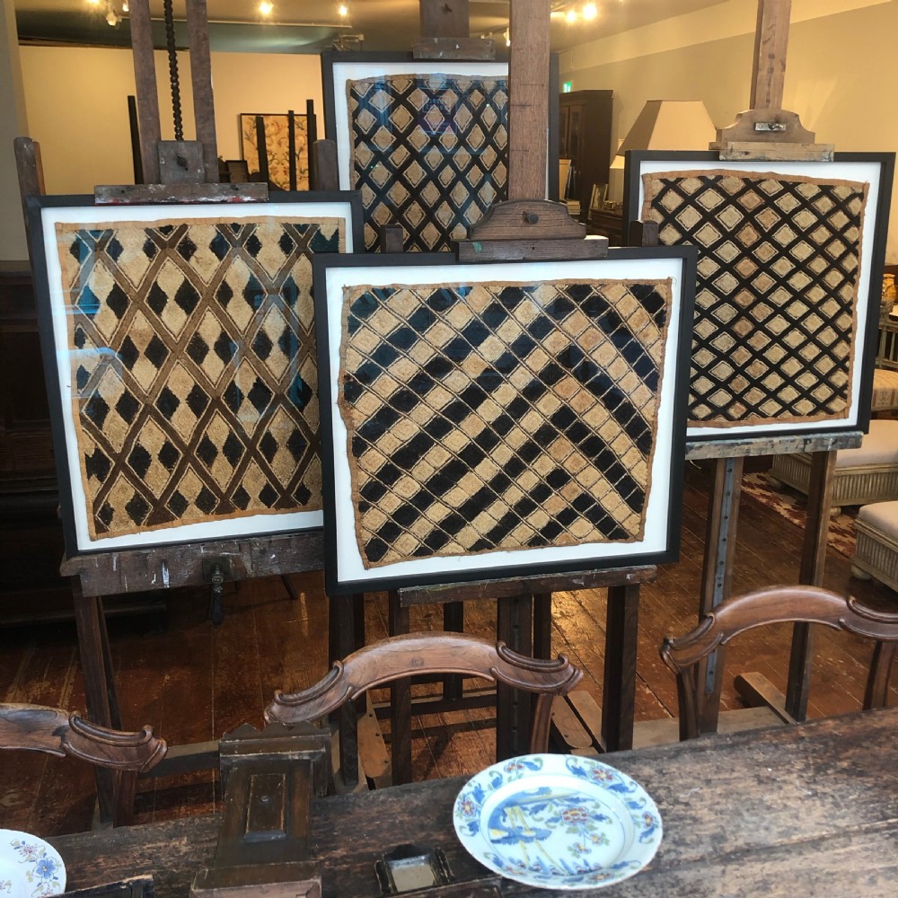 A framed geometric Kuba raffia textile featuring a diagonal grid pattern in natural tan and dark brown tones, presented within a black wooden frame and white mount. View V