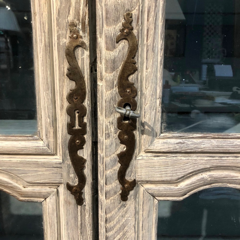 An 18th-century French limed oak vitrine featuring two glazed doors with curved frames, two lower drawers, carved floral detailing, and short cabriole feet. View IV