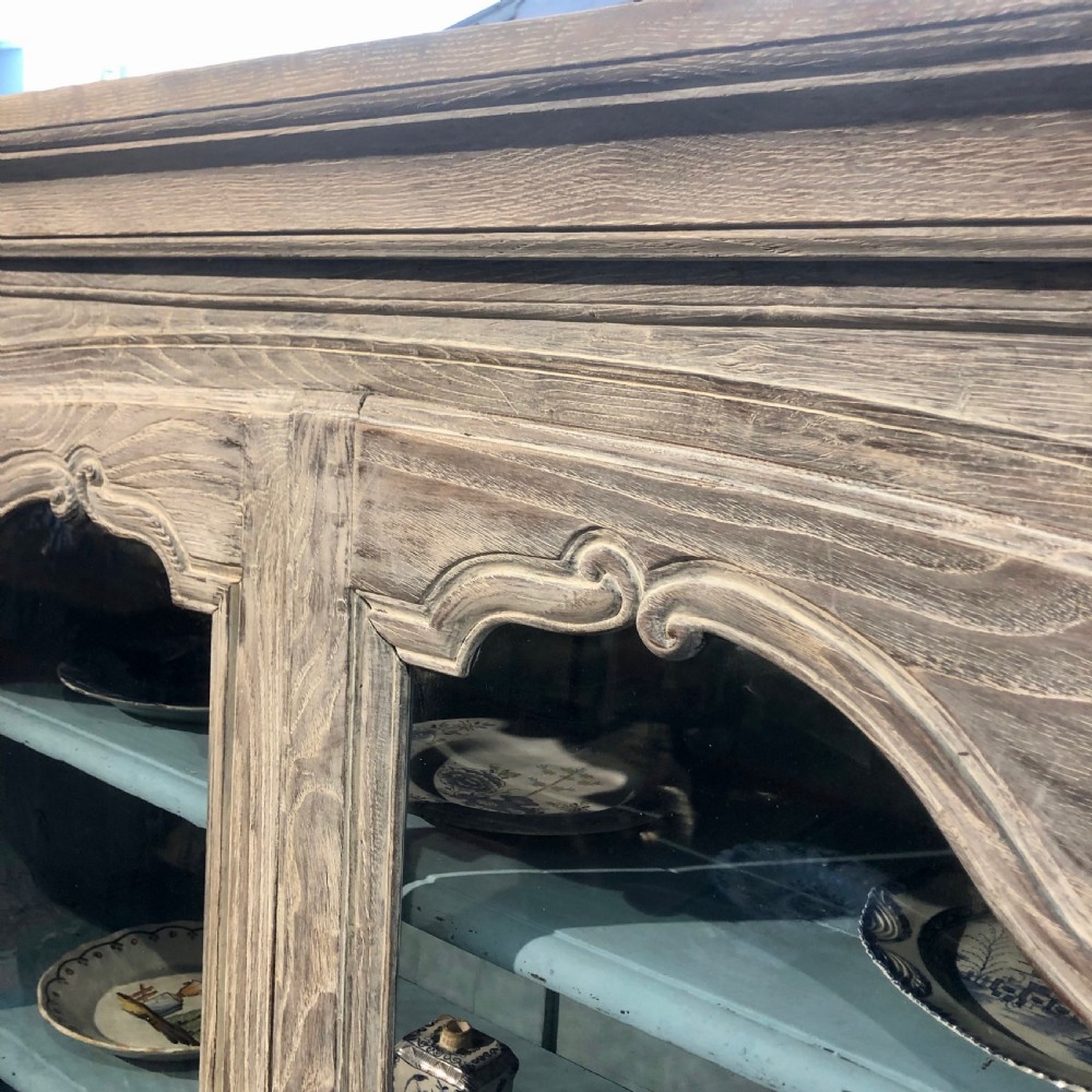 An 18th-century French limed oak vitrine featuring two glazed doors with curved frames, two lower drawers, carved floral detailing, and short cabriole feet. View V