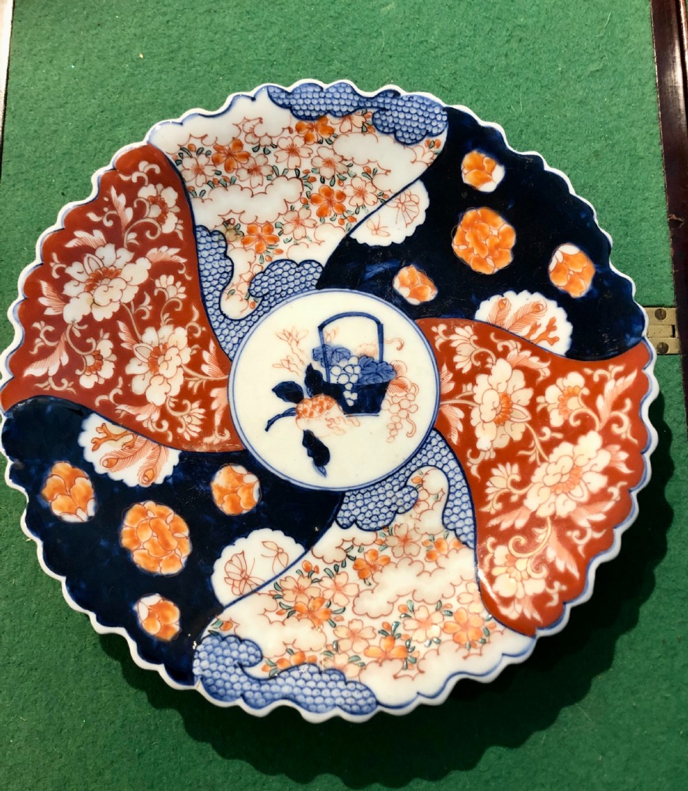 An 18th-century circular Japanese Imari porcelain plate featuring a scalloped rim, cobalt blue and rust red panelled floral patterns, and a central basket motif.