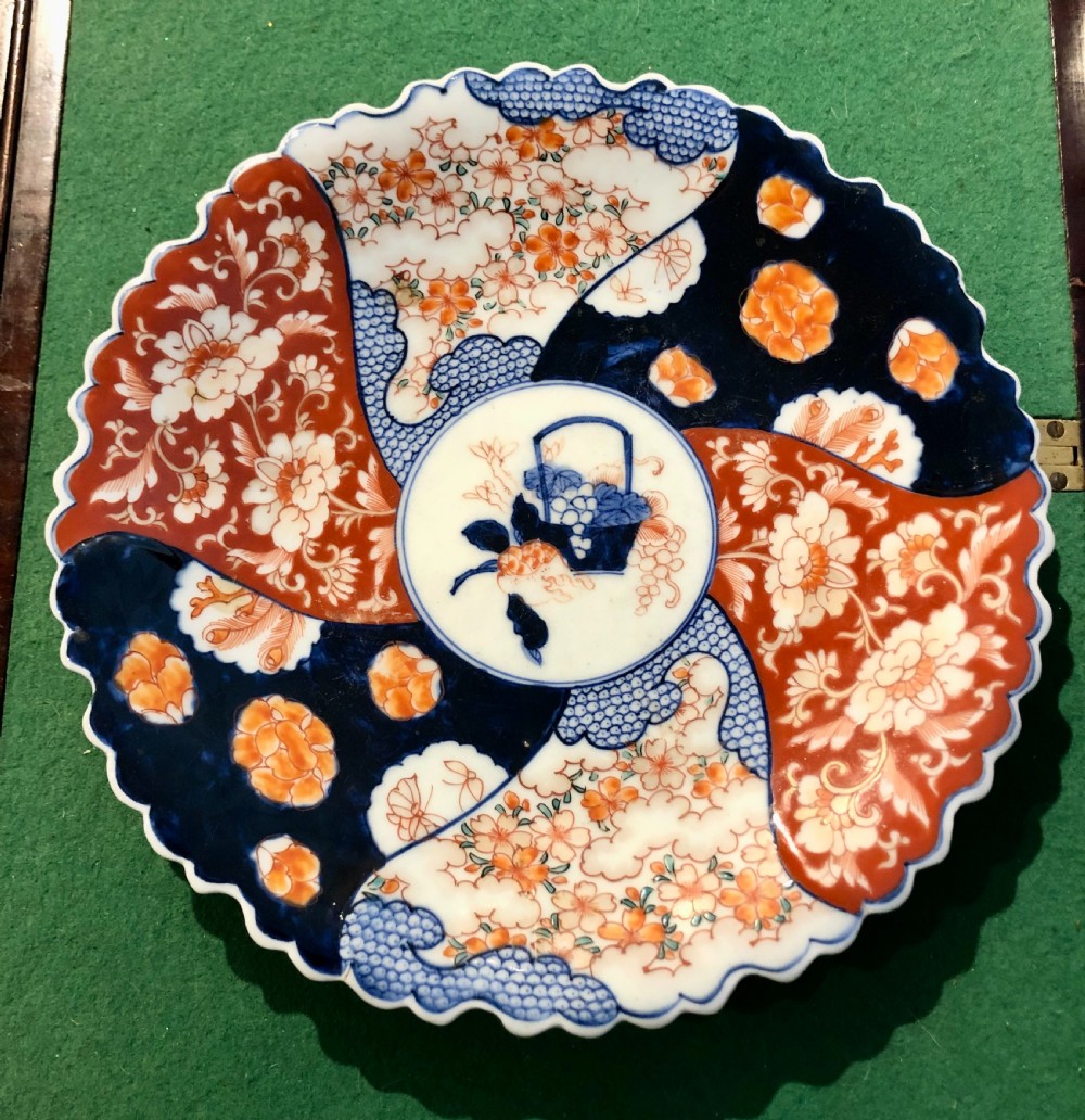 An 18th-century circular Japanese Imari porcelain plate featuring a scalloped rim, cobalt blue and rust red panelled floral patterns, and a central basket motif. View II