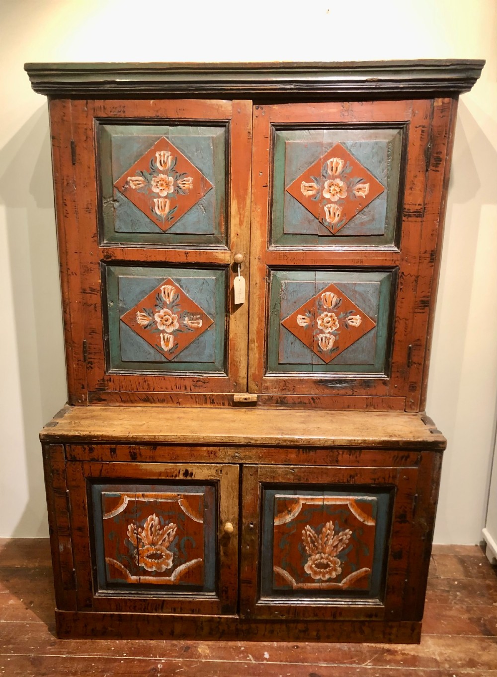 A 19th-century Austro-Hungarian two-tier wooden cupboard decorated with hand-painted floral panels in terracotta red, cream, and teal blue tones.