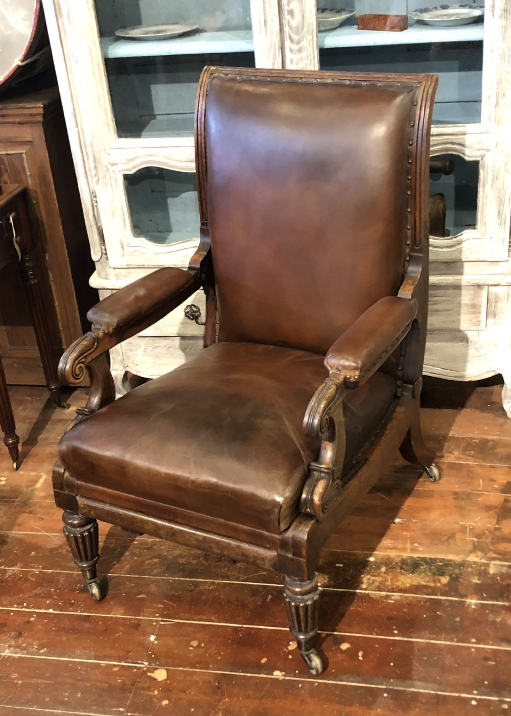 A Regency mahogany library armchair upholstered in warm brown leather with scroll arms, reeded front legs, brass castors, and studding. View X