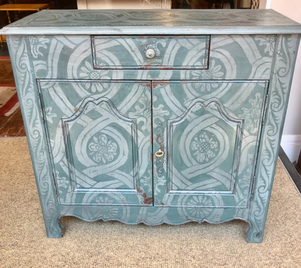 A small wooden cupboard hand-painted in muted sage green with light grey strapwork and floral motifs, featuring a single top drawer and two panelled doors.