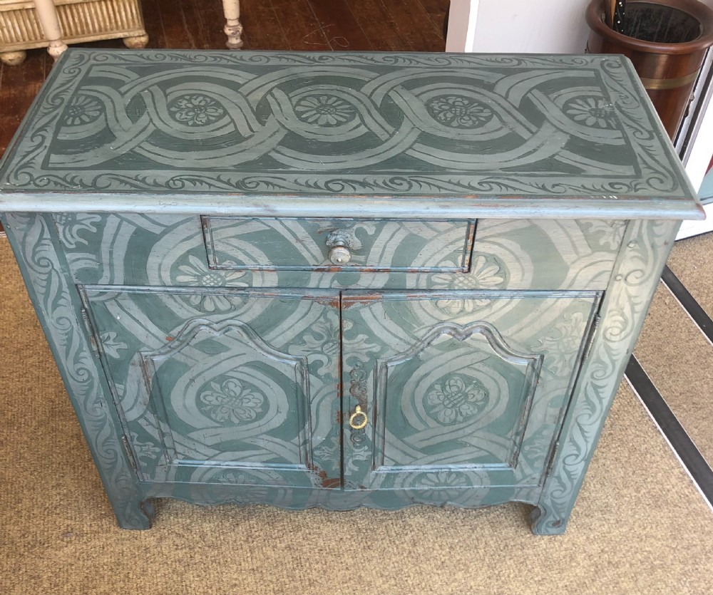 A small wooden cupboard hand-painted in muted sage green with light grey strapwork and floral motifs, featuring a single top drawer and two panelled doors. View III