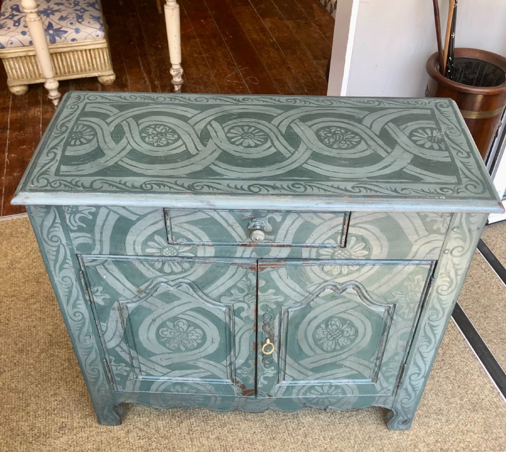 A small wooden cupboard hand-painted in muted sage green with light grey strapwork and floral motifs, featuring a single top drawer and two panelled doors. View IV
